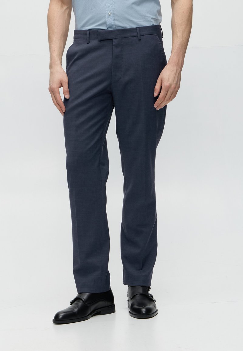 Hombre con pantalones de vestir azul marino, zapatos formales negros y camisa azul claro metida, de pie sobre un fondo blanco liso.
