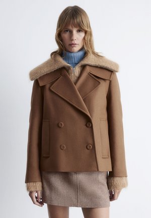 Brauner, doppelt geknöpfter Wollmantel mit einem breiten, weichen Kunstpelzkragen, strukturierten Manschetten und zwei vorderen Taschen. Getragen über einem hellblauen Rollkragenpullover.