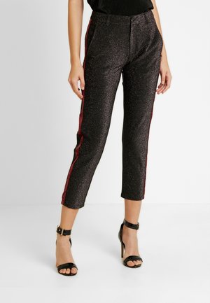 Frau trägt funkelnde schwarze, verkürzte Hose mit burgunderroten Seitenstreifen und schwarze Sandalen mit hohen Absätzen und Knöchelriemen.