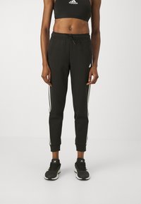 Pantalones deportivos negros con rayas blancas en los laterales, forma ajustada, cinturilla elástica y puños en los tobillos, combinados con zapatillas deportivas negras.