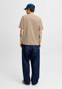 Beige t-shirt med korte ærmer parret med løse blå denim jeans. Synlige tatoveringer på armene, iført en blå cap. Simpel, afslappet pasform.