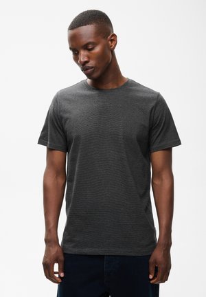 Homme aux cheveux courts portant un t-shirt uni gris foncé à manches courtes, regardant vers le bas avec les mains détendues le long du corps.