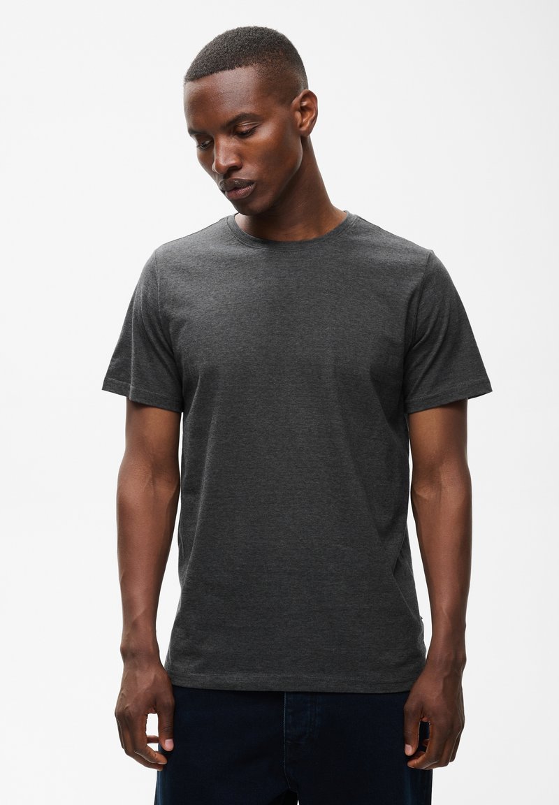 Homme aux cheveux courts portant un t-shirt uni gris foncé à manches courtes, regardant vers le bas avec les mains détendues le long du corps.