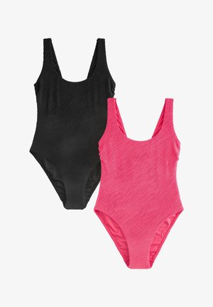 Jeune femme souriante en maillot de bain une pièce rose vif texturé, debout près d’un mur beige avec des volets verts au soleil.