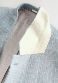 Blazer bleu clair avec un motif rayé subtil, doublure de col crème douce et intérieur gris texturé. Coutures détaillées visibles.
