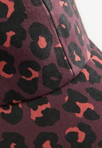 Kepurė su leopardų raštais, tamsiai raudonos fonu, juodomis ir koralo spalvos dėmėmis. Pagaminta iš tekstūruotos medžiagos su siūtomis detalėmis.