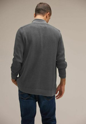Grau gestreifter Cardigan mit Schal-Kragen, langen Ärmeln und detaillierter Strickstruktur, getragen über dunkelblauen Jeans. Subtiles Design mit abgerundetem Saum.
