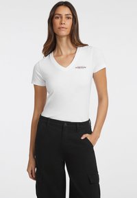Weißes kurzärmeliges V-Neck T-Shirt mit rotem "GUESS JEANS" Logo auf der Brust; kombiniert mit schwarzen Cargo-Hosen. Aus glattem Baumwollmaterial.