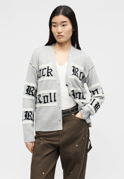 Νεαρή γυναίκα που φοράει γκρι ζακέτα με κείμενο «Rock Roll» πάνω από λευκό πουκάμισο και καφέ παντελόνι, στέκεται με το ένα χέρι στην τσέπη.