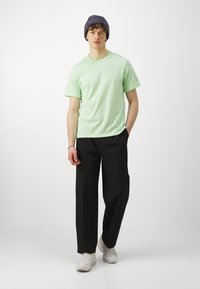 Lysegrøn bomulds t-shirt kombineret med sorte vidde bukser. Modellen har en struktureret marineblå hue og hvide sneakers på.