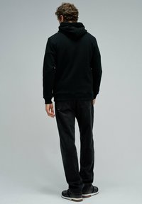 Sudadera negra con capucha de ajuste relajado, combinada con jeans oscuros. La textura de la tela parece suave y lisa. Calzado casual completa el conjunto.