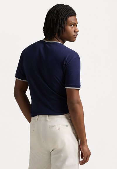 Camisa polo azul-marinho de mangas curtas com detalhes em cinza-claro na gola e nas mangas, combinada com calças em tom creme. Textura de tecido lisa.