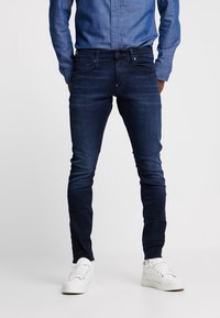 Slim-fit mörkblå jeans med en blekt finish, tillverkade av denim. Har fem fickor och dragkedja. Bärs med en ljusblå skjorta med knäppning.