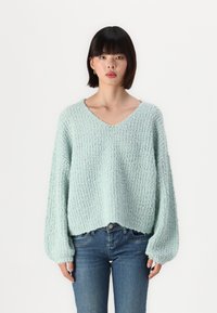 Pull en tricot texturé bleu clair avec un col en V et des manches larges, associé à un jean bleu ajusté. Le pull a une coupe oversize et une texture douce.