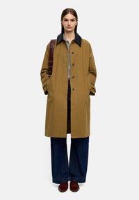 Brązowy, długi płaszcz z kontrastowym czarnym kołnierzem, zapinany na guziki, z bocznymi kieszeniami i luźnym krojem, zestawiony z szerokimi dżinsowymi spodniami i brązowymi butami.