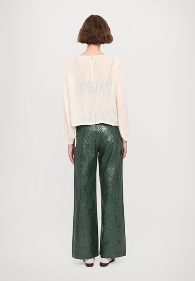 Krämfärgad genomskinlig topp med en texturerad yta, ihop med höga, vida gröna byxor i ett glänsande, skrynkligt material.