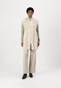 Gilet in faux fur crema con colletto alto e zip frontale, indossato sopra una maglietta verde chiaro e abbinato a pantaloni beige a gamba larga e stivali neri.
