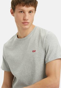 Giovane uomo che indossa una t-shirt grigia chiaro di Levi's con logo rosso, guarda leggermente di lato su uno sfondo bianco semplice.