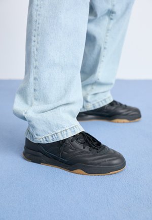 Zapatillas de cuero negro con una superficie texturizada, que cuentan con una suela de goma y cordones negros. Combinadas con unos jeans de mezclilla azul claro.