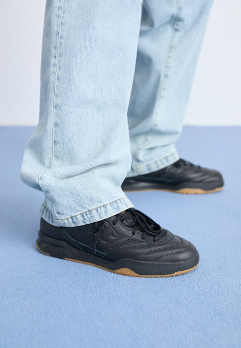 Zapatillas de cuero negro con una superficie texturizada, que cuentan con una suela de goma y cordones negros. Combinadas con unos jeans de mezclilla azul claro.