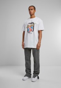 Hvid bomulds t-shirt med et farverigt grafisk design på fronten, parret med grå denimbukser og hvide sneakers.