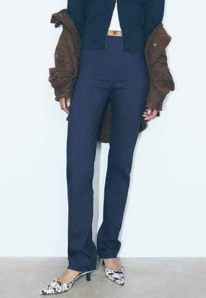 Marinblå högmidjade byxor med en strömlinjeformad passform. Kombinerade med en brun jacka och mönstrade spetsiga klackskor. Texturerat tyg synligt.
