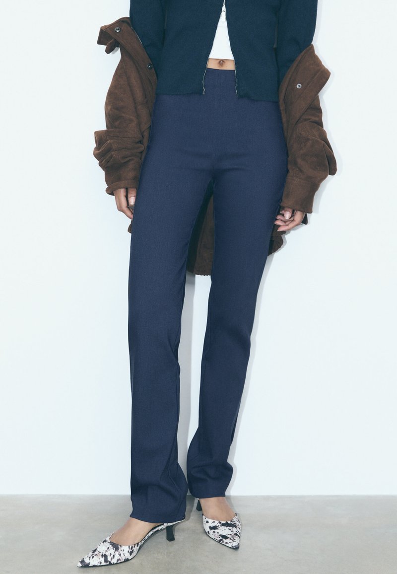 Tmavě modré kalhoty s vysokým pasem a výrazně přiléhajícím střihem. Kombinovány s hnědým sáčkem a lodičkami s vzorovanou špičkou. Viditelná texturovaná látka.