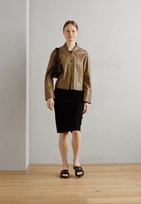 Brun lærjakke, svart tettsittende skjørt, svarte slippers med spenner, minimalistisk håndveske, glatte teksturer og nøytral bakgrunn.