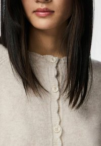 Kremowy sweter rozpinany na guziki z karbowanym dekoltem i pięcioma okrągłymi guzikami. Miękka tekstura i widoczne długie, proste ciemne włosy.