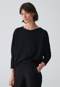 Schwarzer, locker sitzender Pullover mit rundem Halsausschnitt und langen Ärmeln, kombiniert mit hochgeschnittenen schwarzen Hosen mit Knopfdetails.