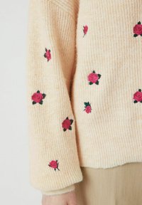 Pletena majica svijetlo b breskve s rebrastom teksturom. Sadrži male, vezene ružičaste ruže s zelenim listovima širom tkanine.