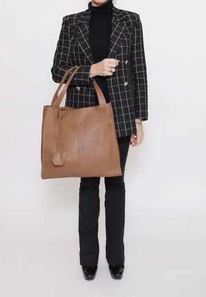 Borsa tote in pelle marrone con due manici lunghi, texture liscia e un ciondolo esagonale. Indossata con blazer nero a quadri e pantaloni neri.