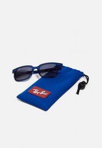 Mėlynos plastikinės saulės akinius su tamsiais lęšiais, derinamos su atitinkamu mėlynu raišteliu maišeliu, ant kurio yra raudonas Ray-Ban logotipas.