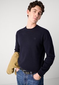Navy blue sweater with a round neck and ribbed cuffs, featuring a small, embroidered logo on the chest. Paired with light denim jeans.