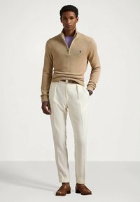 Bež pulover na zadrgo z rebrastimi zapestnimi manšetami, kombiniran s kremnimi hlačami z naborki in rjavi usnjeni čevlji, stoječi ob nevtralnem ozadju.