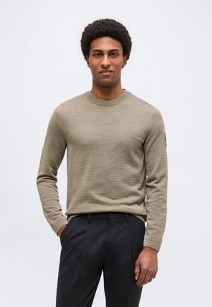 Άνδρας με κοντά σγουρά μαλλιά, φοράει μπεζ πουλόβερ με μακριά μανίκια και μαύρο παντελόνι, στέκεται με το ένα χέρι στην τσέπη.