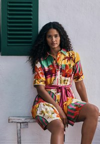 Femme aux longs cheveux bouclés assise sur un banc en bois, portant une robe imprimée tropicale colorée avec une ceinture rose, contre un mur blanc avec des volets verts.
