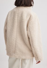 Vrouw in een crèmekleurige oversized sherpa fleece jack en lichte geribde broek, vanuit de rug weergegeven tegen een effen achtergrond.