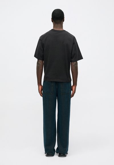 Juodas medvilnės marškinėliai su trumpomis rankovėmis, derinami su laisvai krentančiais tamsiai mėlynais džinsais, turinčiais tiesų kirpimą ir dviem galiniais kišenėmis.