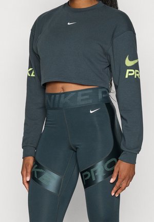 Osoba nosząca ciemnozieloną, krótką bluzę Nike Pro oraz legginsy z wysokim stanem z logo Nike na rękawach, piersi i udach.