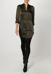 Abito in satin verde oliva con scollatura a V, vita arricciata e maniche lunghe. Indossato con collant neri e stivaletti alla caviglia con accenti di fibbia.