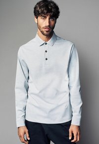 Șir de polo lung de culoare gri deschis, cu trei nasturi negri, guler tradițional și un finisaj texturat, asortat cu pantaloni întunecați.