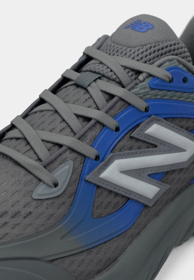 Zapatilla deportiva gris con parte superior de malla, acentos azules y logo plateado reflectante. Presenta cordones grises y suela acolchada para mayor soporte.