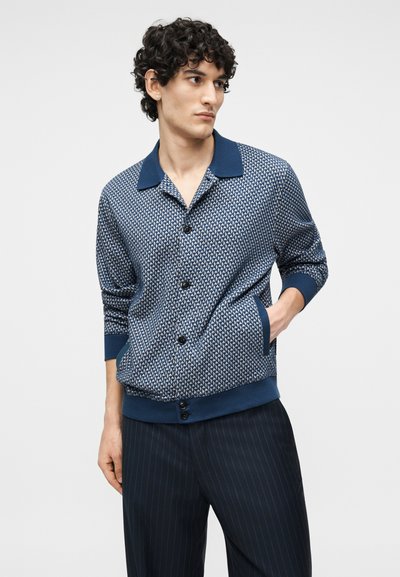 Άνδρας που φοράει μπλε πουκαμισοειδή ζακέτα με τσέπες και σκούρο παντελόνι με λεπτές ρίγες, όρθιος με το ένα χέρι στην τσέπη.