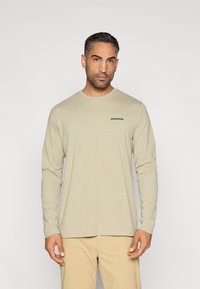 Πουκάμισο με μακριά μανίκια σε ανοιχτό μπεζ ύφασμα, με στρογγυλή λαιμόκοψη και μικρό μαύρο λογότυπο "Patagonia" στη αριστερή πλευρά του στήθους.