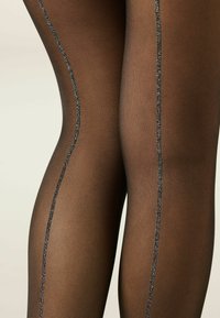 Durchsichtige schwarze Strumpfhosen mit einer dezenten gerippten Textur und einem glänzenden schwarzen glitzernden Streifen, der vertikal entlang des äußeren Beins verläuft.