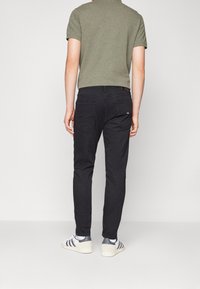 Hombre con camisa de manga corta verde oliva, jeans ajustados negros y zapatillas Adidas blancas de pie de espaldas a la cámara.