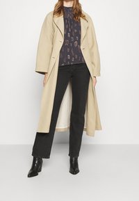 Cappotto lungo beige con rever, abbinato a una maglia navy a fantasia e jeans neri a gamba dritta. Stivali neri con tacco completano l'outfit.
