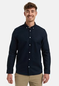 Hymyilevä nuori mies, joka pukeutunut tummansiniseen nappipaitaan ja beigenvärisiin housuihin, seisoo yksinkertaisen valkoisen taustan edessä.