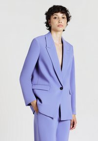 Persona con capelli corti e ricci che indossa un completo su misura di un leggero colore viola con un bottone allacciato, in piedi contro uno sfondo bianco uniforme.